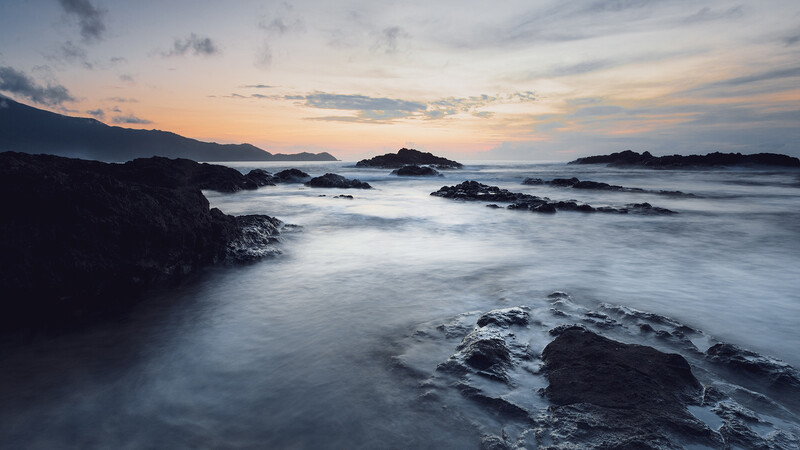 Rocky coastline at twilight with smooth water flowing between dark boulders and distant islands.