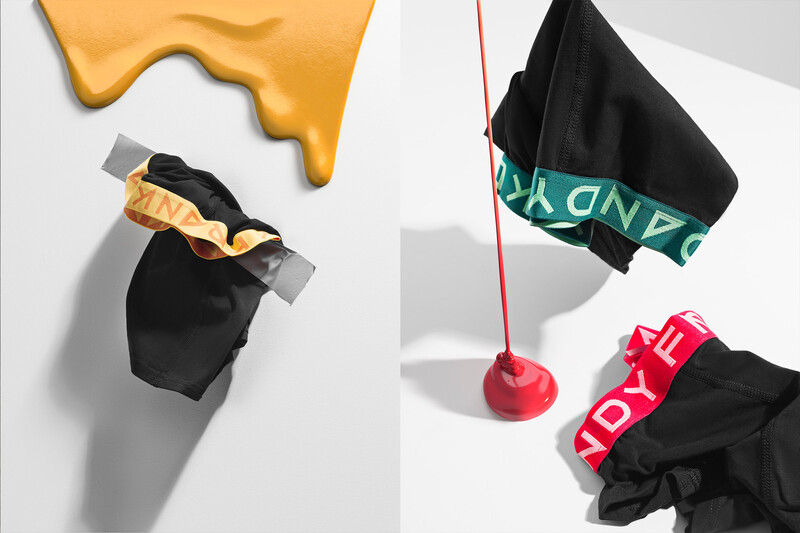 Flat lay arrangement of black clothing items with colorful accessories and a yellow abstract shape against white background.