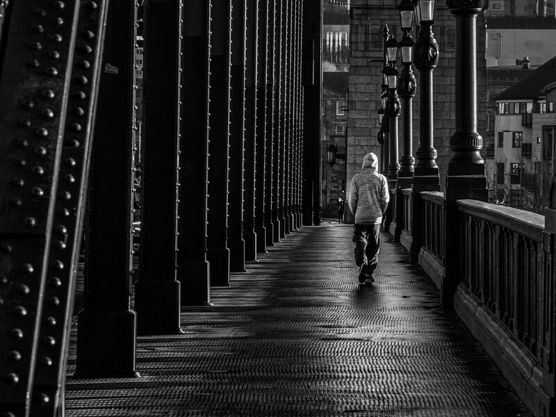 Solitary figure jogging across an industrial metal bridge with strong linear perspective and dramatic shadows.