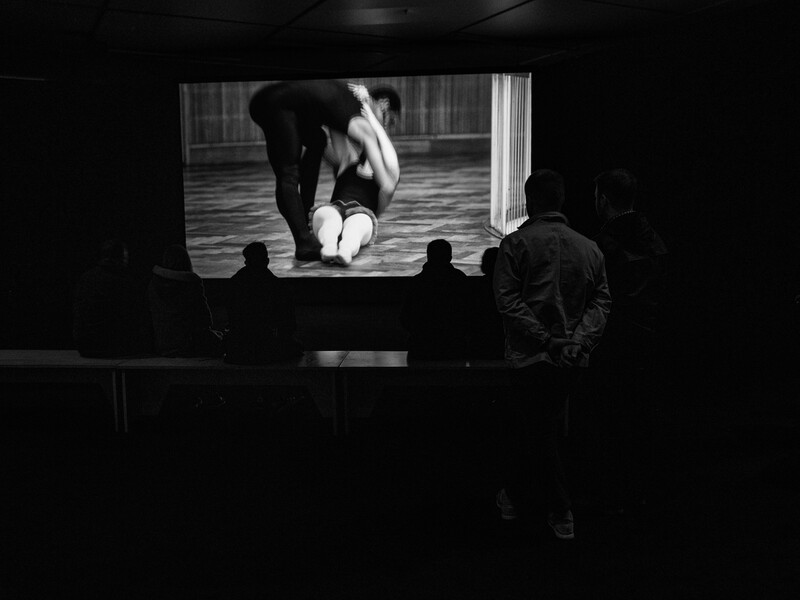 Silhouetted audience members watching a black and white film projected on a screen showing a rhinoceros.