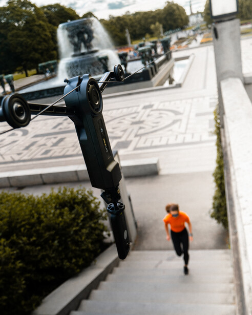 Skateboarder performing aerial trick above concrete plaza with spectator in orange shirt watching from pathway below.