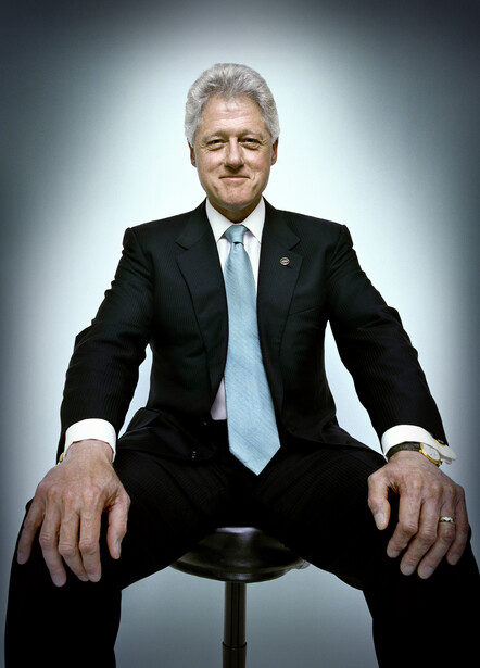 Older man in dark suit and light blue tie seated on stool against gradient background.