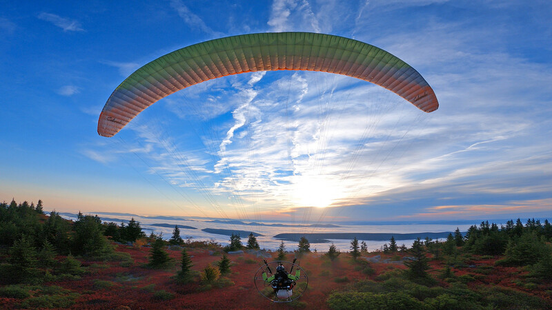 Paraglider in flight above forested terrain with a lake and sunset sky in the background.
