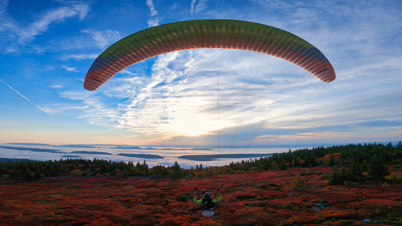 Paraglider in flight above a red-toned landscape with forested ridges and a lake at sunset.