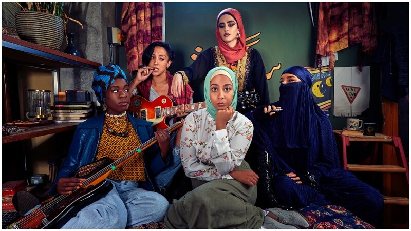 Five women of diverse backgrounds posing together in a music studio with instruments and warm interior lighting.