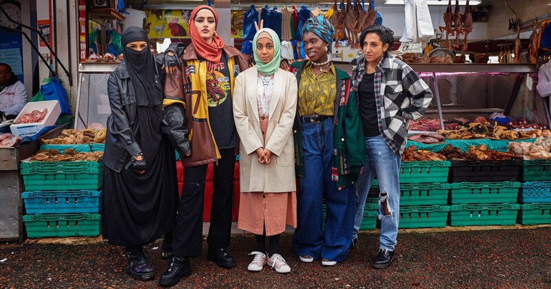 Five women in diverse cultural attire standing together at an indoor market with green crates of produce behind them.
