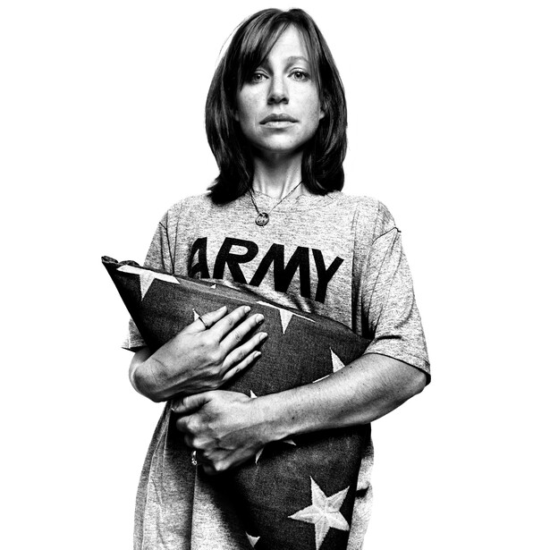 Black and white portrait of a woman holding a book, wearing an army-print t-shirt, looking directly at camera.