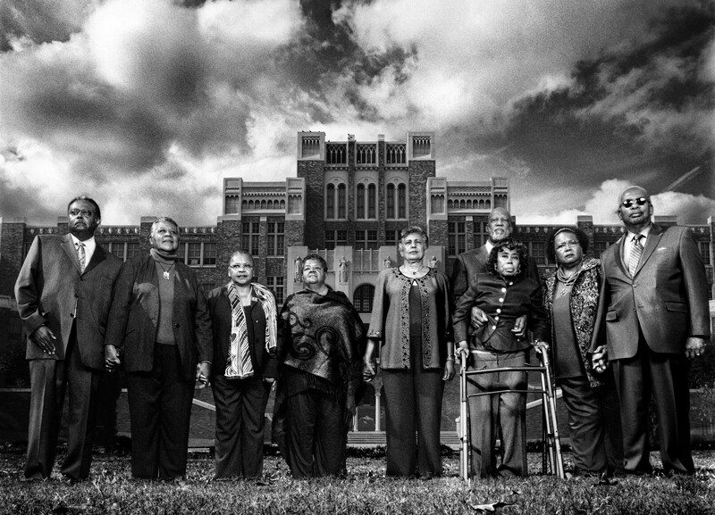 Group of ten people in formal attire standing in a line before a brick building with dramatic cloudy sky.