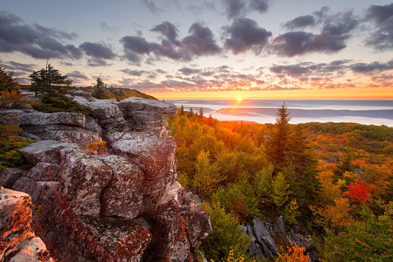 Rocky outcrop in foreground with autumn forest and glowing sunset over distant water.