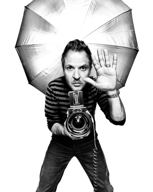 Photographer holding large format camera pointed at viewer, positioned under illuminated umbrella reflector.