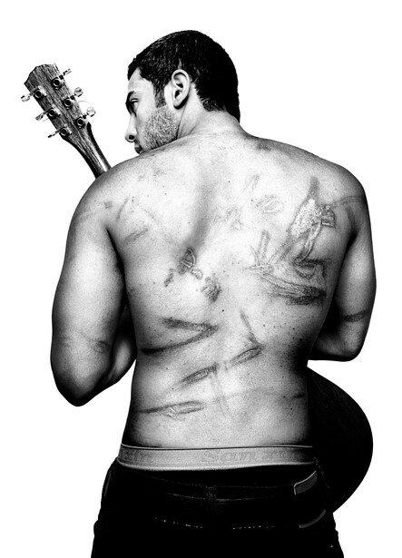 Shirtless man with visible tattoos on his back and torso, holding an acoustic guitar over his shoulder.