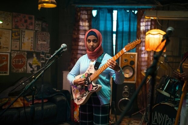 Musician wearing red hijab and sunglasses playing electric guitar in a studio setting.