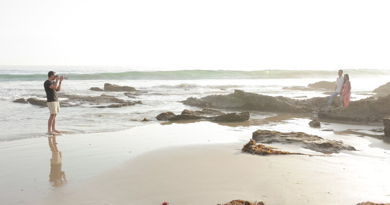 Photographer standing in shallow water capturing images of people on a rocky beach during golden hour.