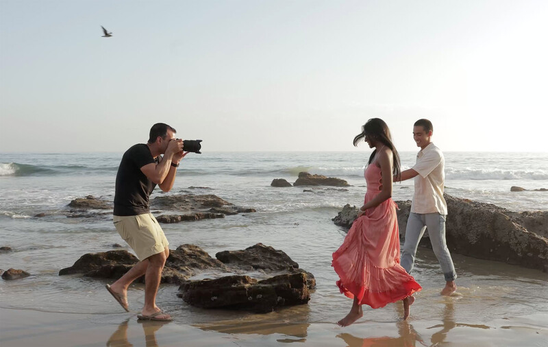 Photographer taking a portrait of two people in shallow ocean water with rocky outcrops.