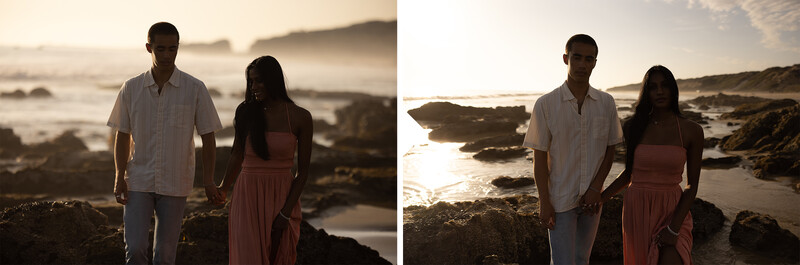Couple holding hands on rocky coastline at golden hour with calm ocean and distant hills.