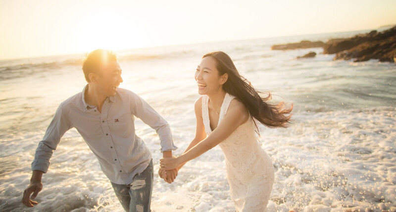 Couple holding hands while running through shallow ocean waves at sunset.