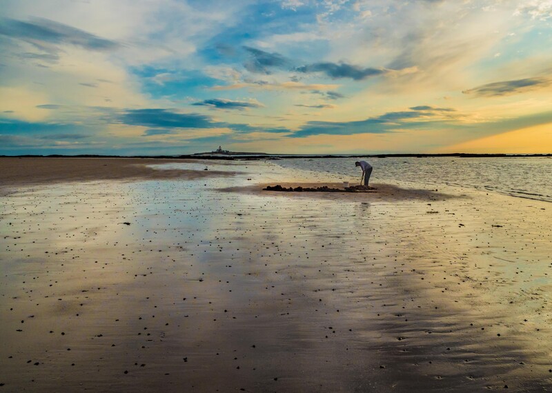 Figure standing in shallow water on an expansive tidal flat at sunset, with dramatic sky above.