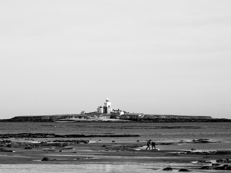Military warship anchored in shallow water near a rocky shoreline, viewed from a beach with figures in the foreground.