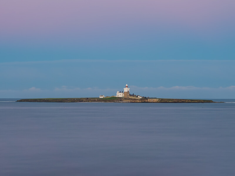 A small fortified island with a lighthouse and stone structures sits in still blue water under a twilight sky.