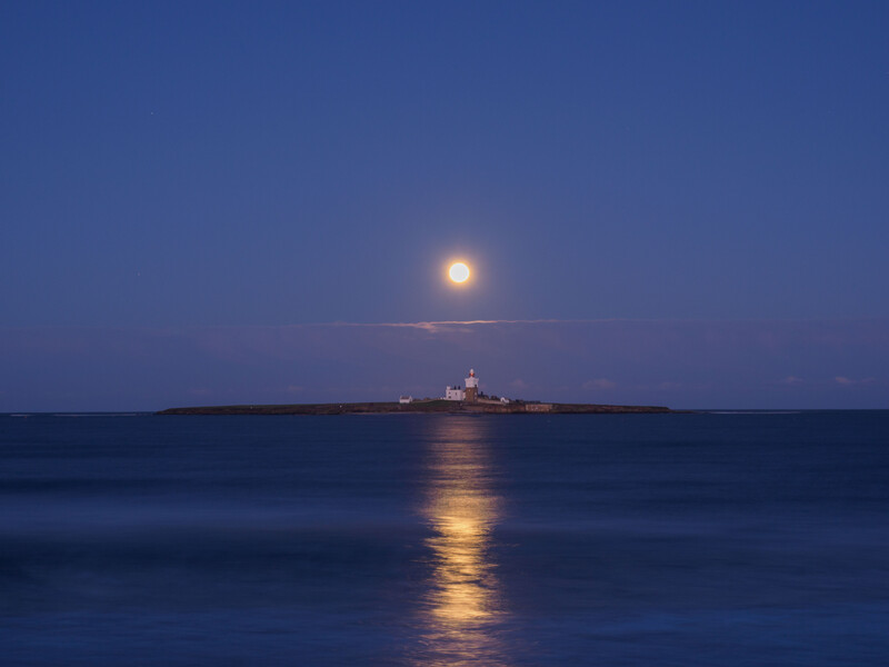Full moon rising above a distant lighthouse silhouetted on the horizon at twilight.