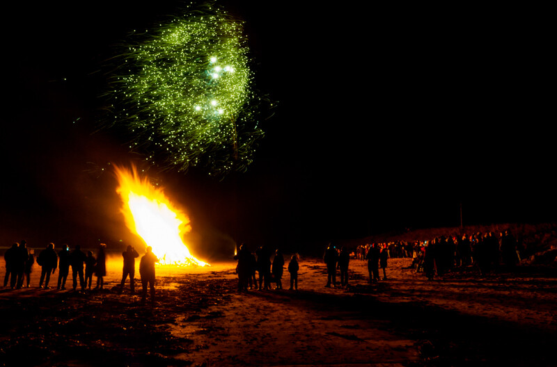 Large bonfire erupting with bright yellow and orange flames while green and yellow sparks explode upward into the dark sky, surrounded by silhouetted spectators on a beach.