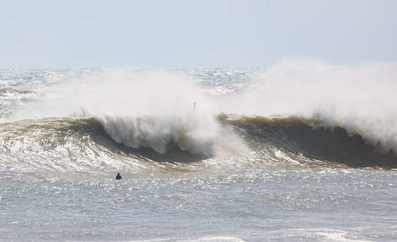 Large ocean waves with white foam crashing toward shore, a surfer visible in the water.