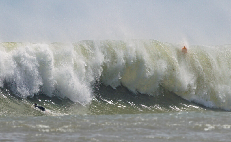 Massive ocean wave with white foam crest rolling toward shore under overcast sky.
