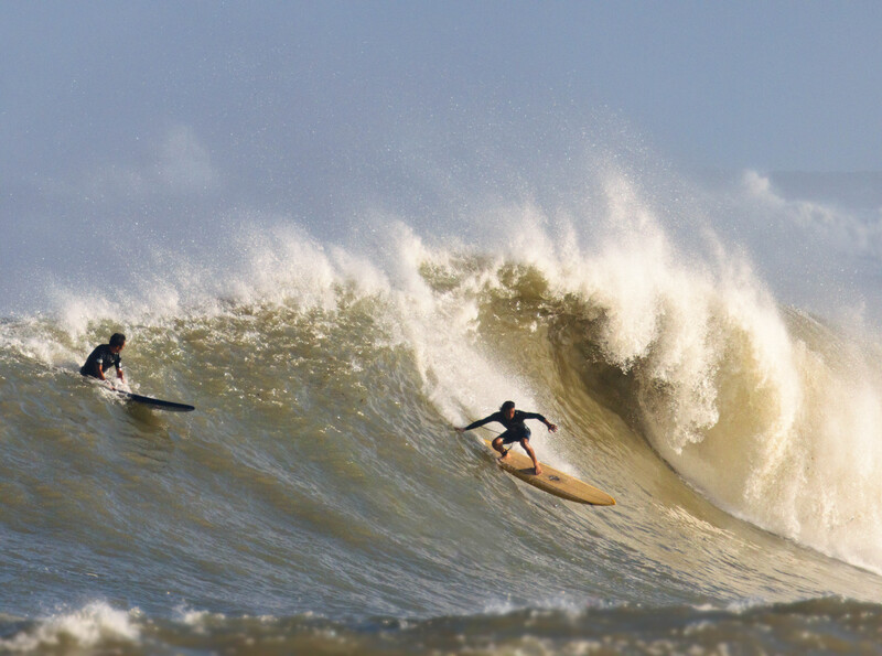 Two surfers riding a massive wave with white foam spray and dramatic backlighting.