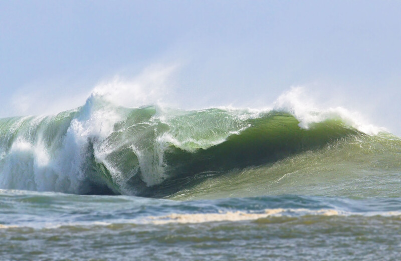 Massive ocean wave with white foam crest and translucent green face breaking near shore.