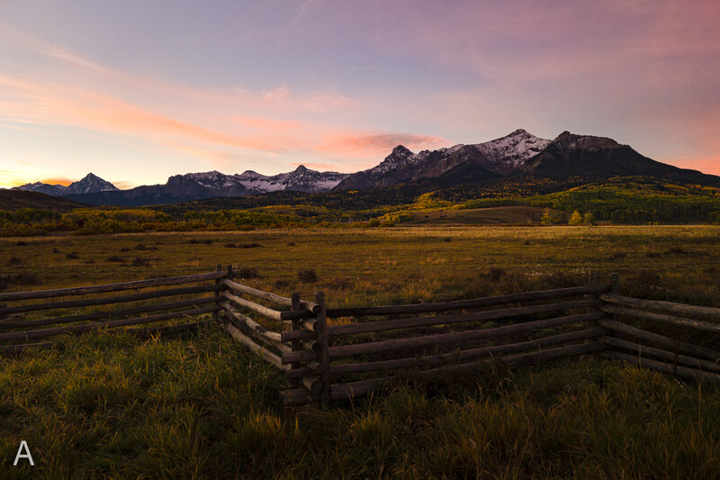 Rustic wooden ranch fence in foreground with snow-capped mountains at sunset.