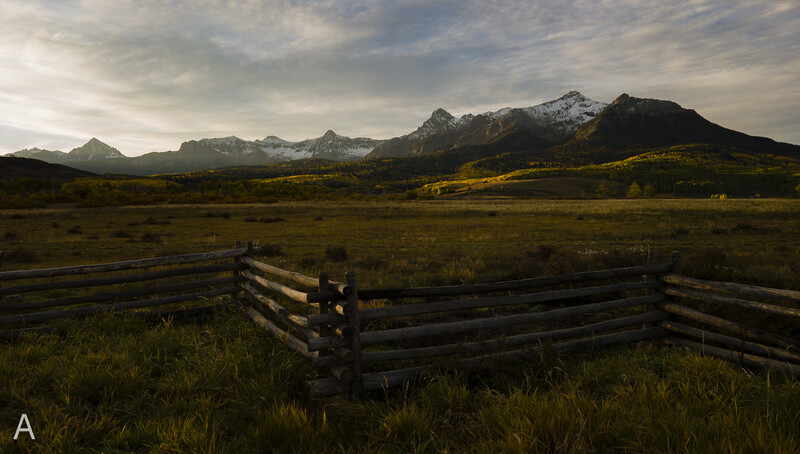 Weathered wooden fence in a grassy meadow with snow-capped mountains under dramatic cloudy sky.