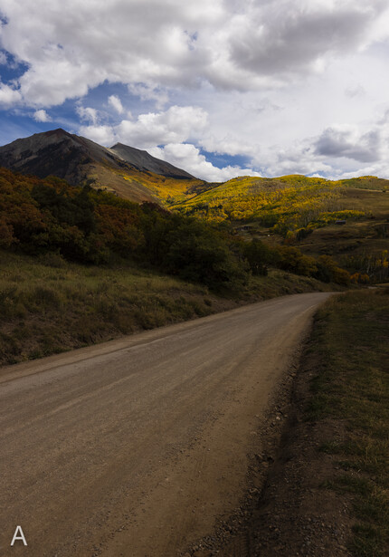 Unpaved road stretching through mountainous terrain with golden hillside vegetation and dramatic cloudy sky.
