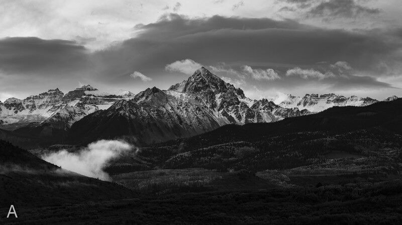 Snow-capped mountain peak rising above forested valleys with dramatic storm clouds overhead in black and white.