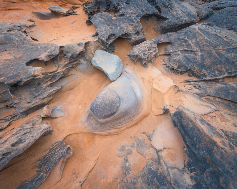 Smooth, rounded rock formation surrounded by layered stone and rust-colored sand in a desert landscape.