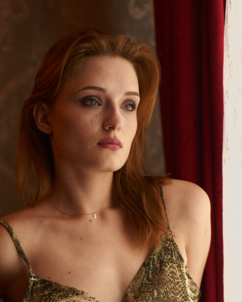 Woman with reddish-brown hair wearing a gold sequined top, posed against a dark background with red curtain.