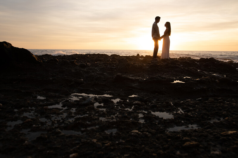 Silhouetted couple standing on rocky shoreline at sunset with golden sky and distant cityscape.