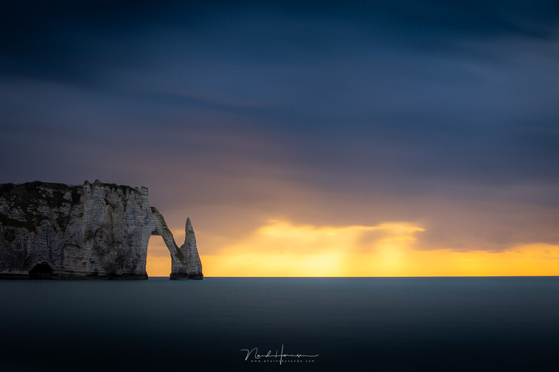 Dramatic coastal rock formation silhouetted against a golden sunset over calm water.