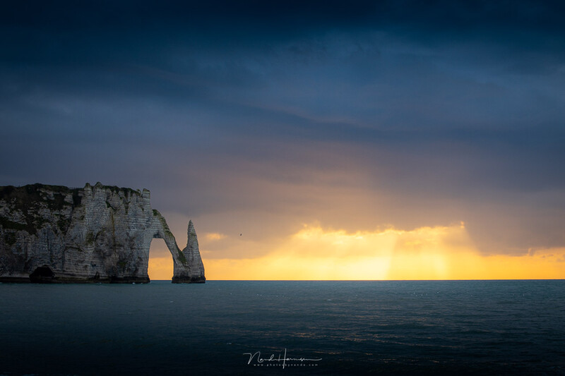 Dramatic coastal rock formation with natural arch silhouetted against golden sunset over calm ocean.
