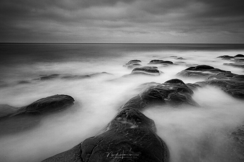 Long-exposure black and white seascape with smooth water flowing around dark rocks under stormy skies.