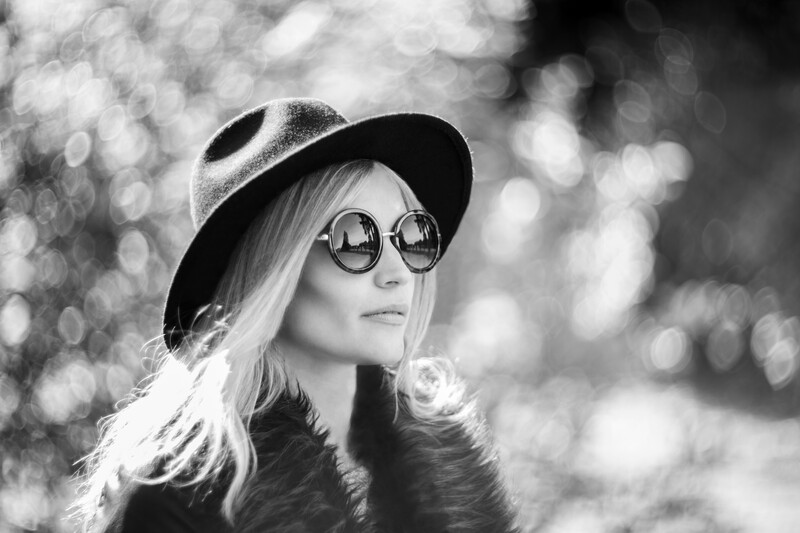 Woman wearing round sunglasses and a wide-brimmed hat, photographed in black and white with soft bokeh background.