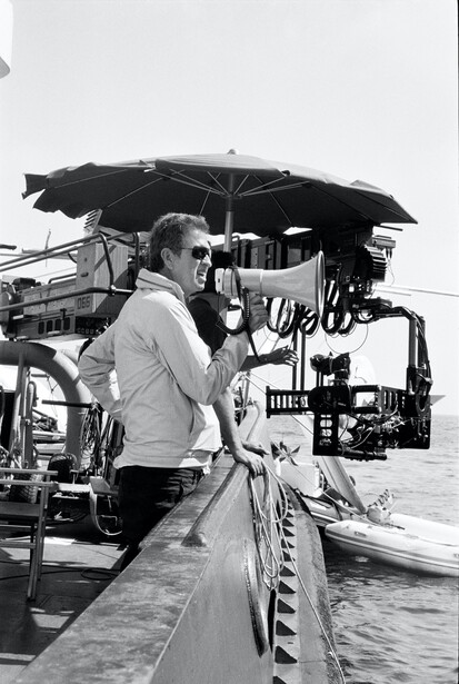 Photographer with camera on boat deck holding rope, black umbrella overhead for sun protection.