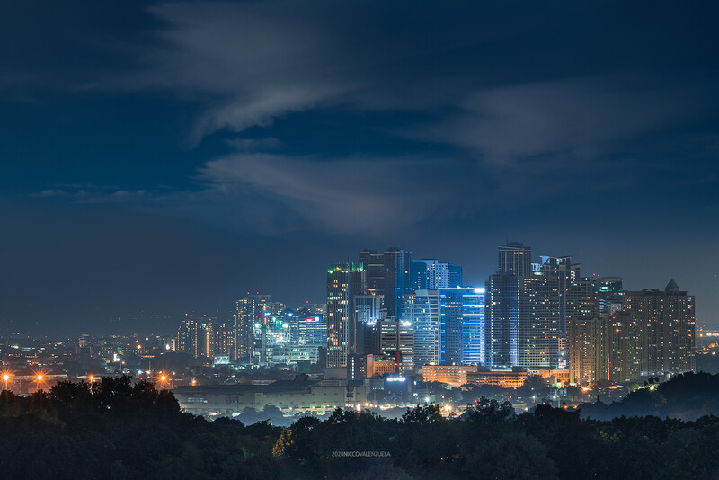 Illuminated downtown skyline at dusk with tall buildings reflected in water, forested hills in foreground.