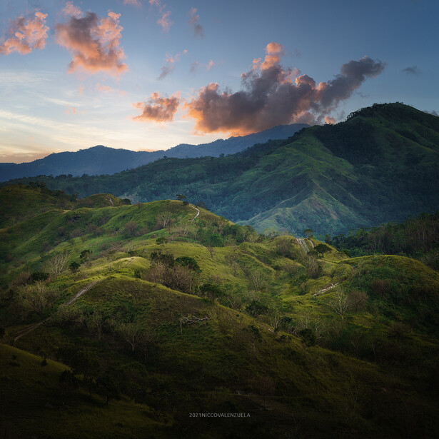 Rolling green hillsides with dramatic orange-tinted clouds at sunset and mountains in the distance.