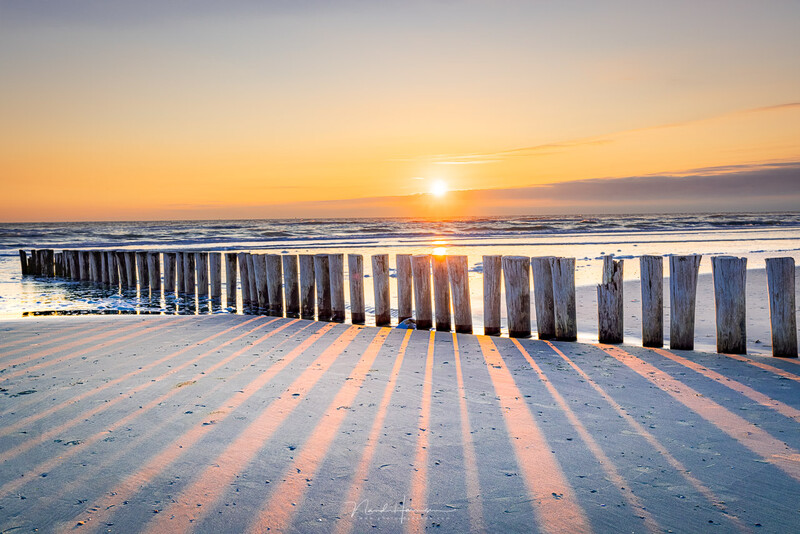 Wooden groynes extending into the sea at golden hour sunrise with shadows cast across the beach sand.