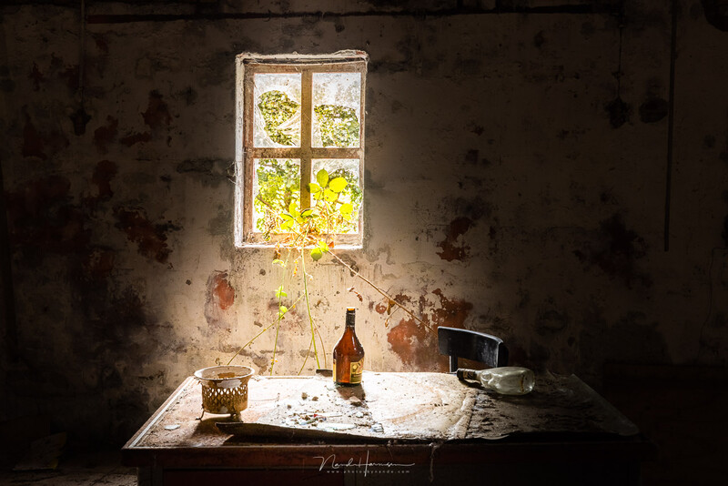 Abandoned interior room with deteriorating walls, a single window with bright natural light, and a table displaying a bottle, glass, and scattered objects.