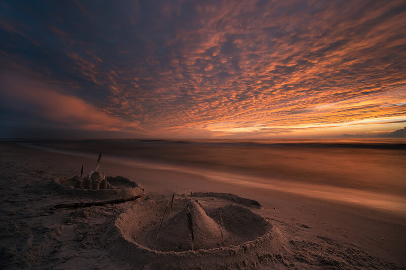 Dramatic sunset sky with orange and gold clouds over a beach with sandcastle structures in foreground.