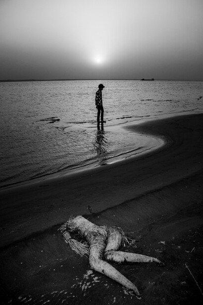 Silhouetted figure standing in shallow water at sunset, with another figure lying on the beach in the foreground.