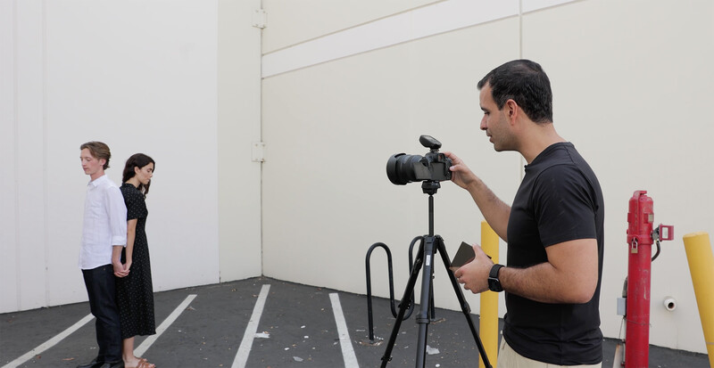 Photographer on tripod shooting two subjects in a studio with white walls and marked floor.