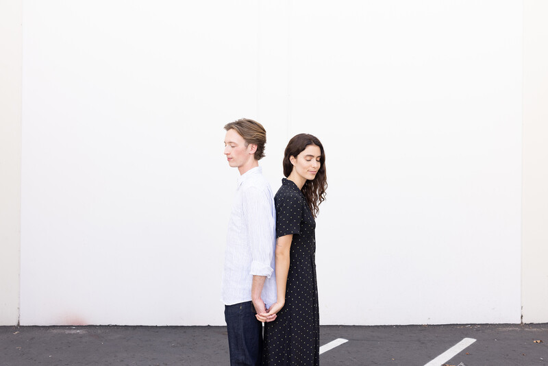 Two people holding hands standing against a white wall with gray pavement below.