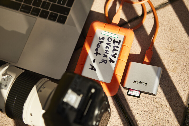 Overhead view of photography workspace with camera, laptop, lens, and orange color reference card on carpet.